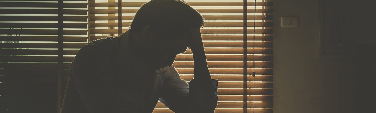 A silhouette of a man sitting on his bed with his hand against his forehead, seemingly upset, light seeping from the blinds.
