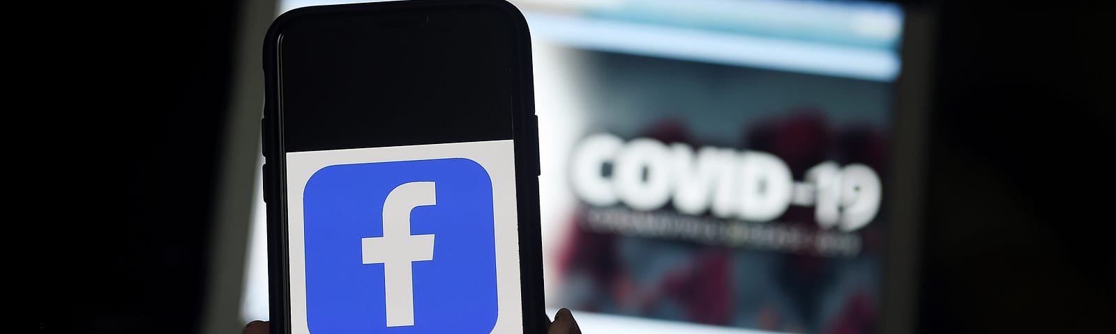 Facebook logo is displayed on a mobile phone screen with the words COVID-19 blurred in the background on a laptop screen