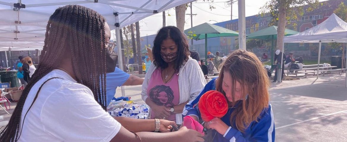 Teen girl with box braids wearing mask and white T-shirt handing kit to another teen girl wearing a blue jacket across a table on a sunny day.