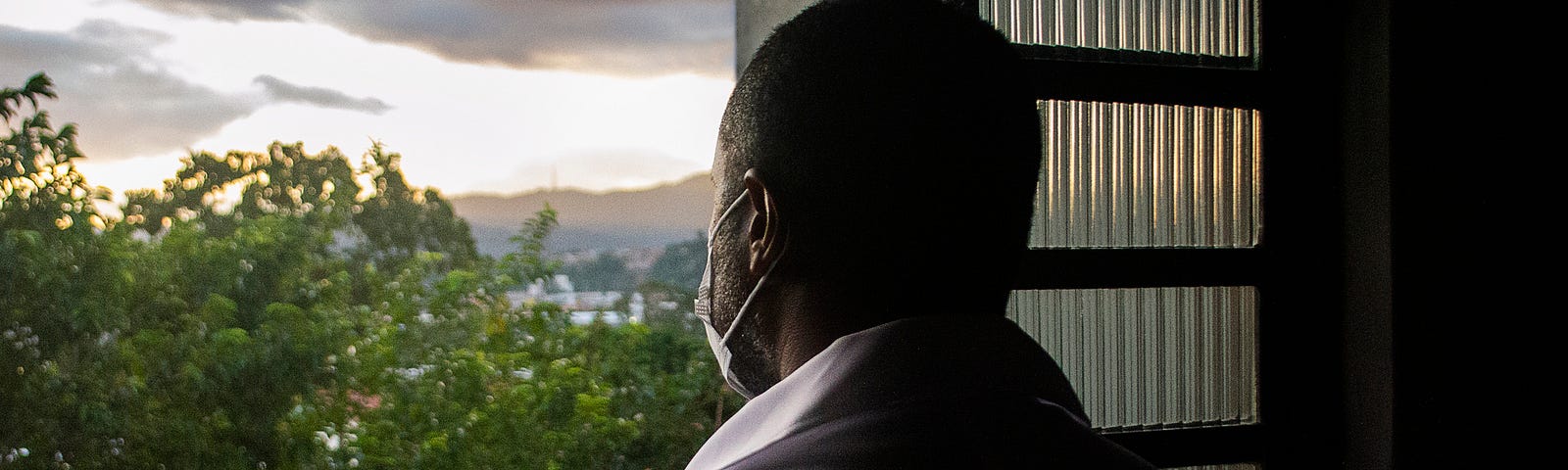 A photo from the back of a black man wearing a face mask looking out his window.