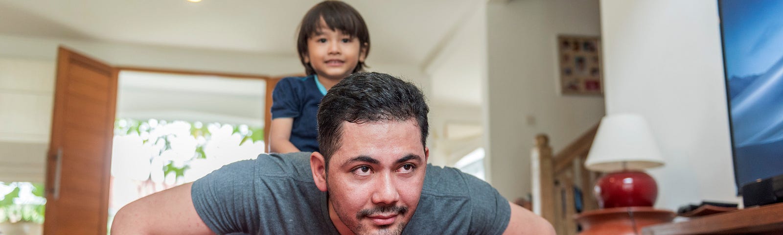 A photo of a father doing push ups in the living room while his son sits on his back.