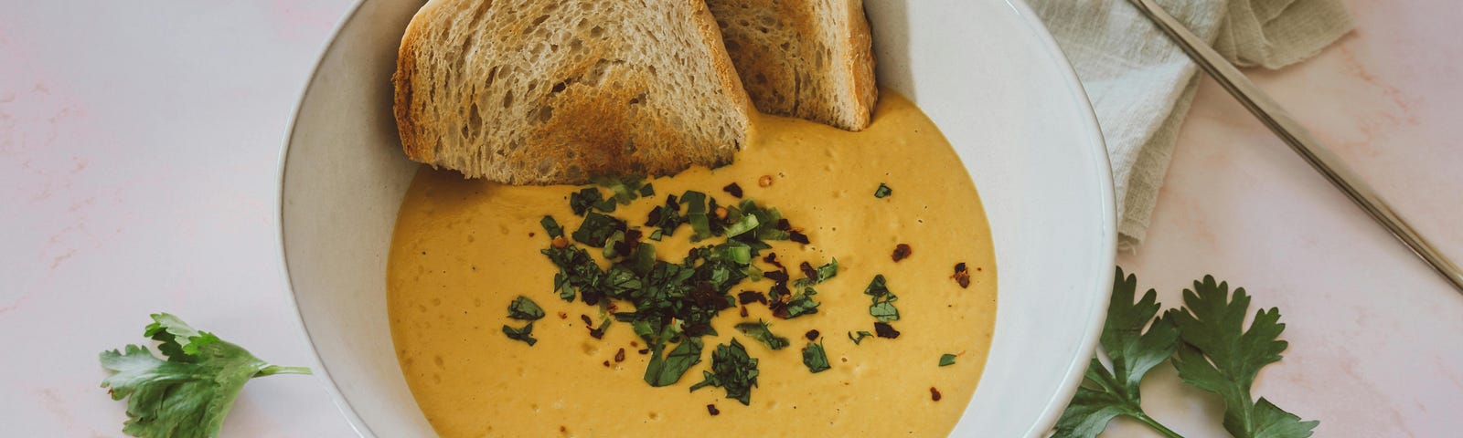 A bowl of creamy yellow soup garnished with chopped fresh parsley sits on a light-colored surface. Two toasted white bread rest on the edge of the white bowl, and a silver spoon lies nearby on a linen napkin.