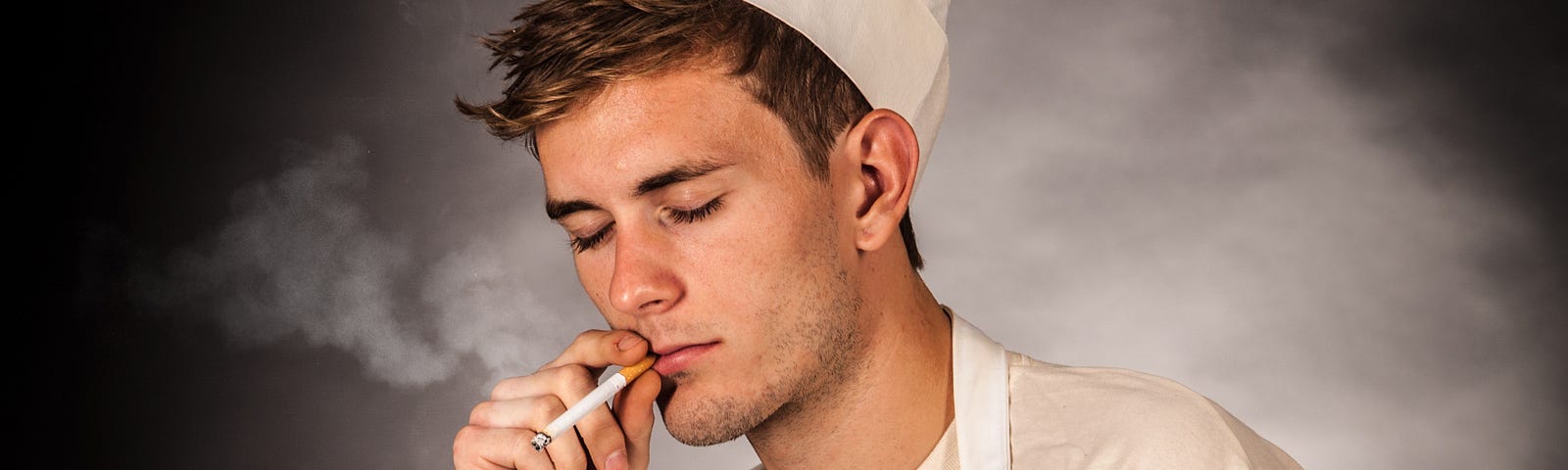 A male cook with a chef’s cap smoking a cigarette.
