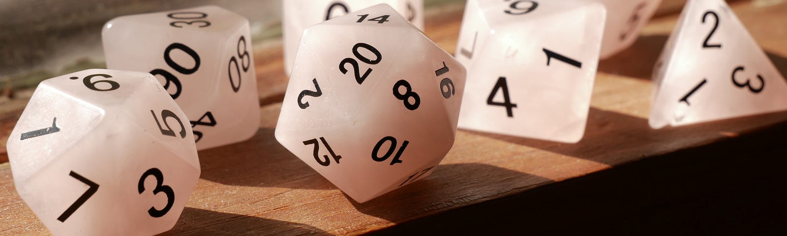 On a wooden window sill, there is a variety of white dice that range from a D4 to a D20. They have white faces with black numbers.