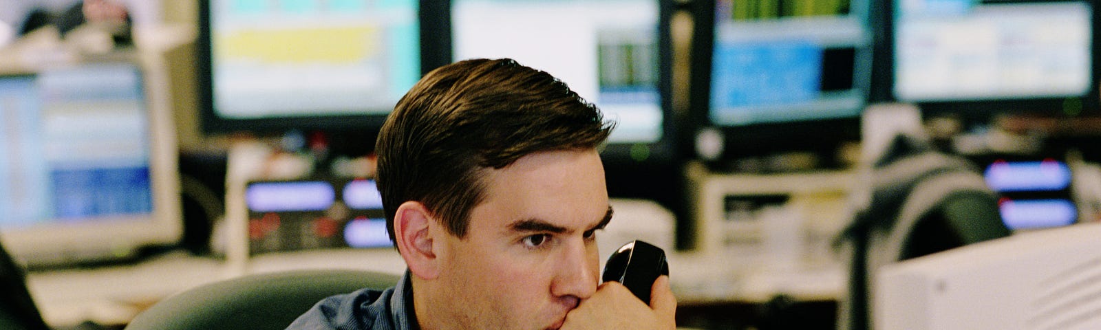 A broker contemplating and holding up a phone while watching his computer.