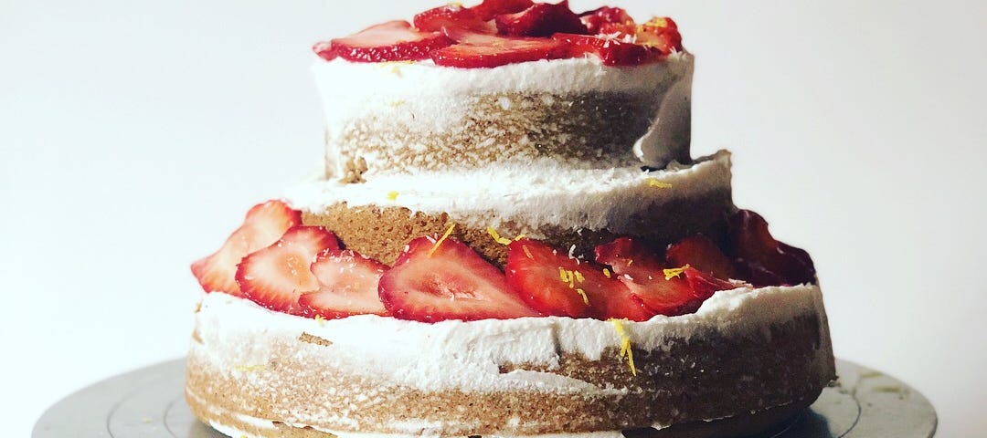 A layered cake on a silver cake stand — the cake is golden brown with lightly applied white icing and strawberries.