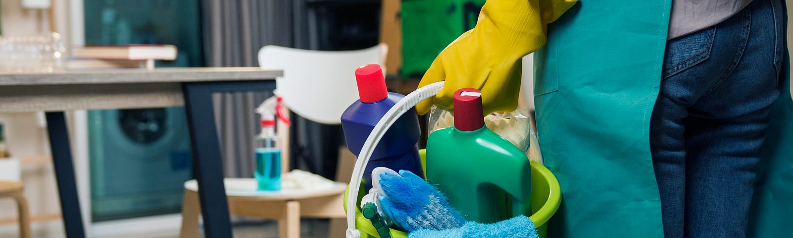 A waist-down shot of a maid, seen from behind, holding a bucket of cleaning products in a small room.