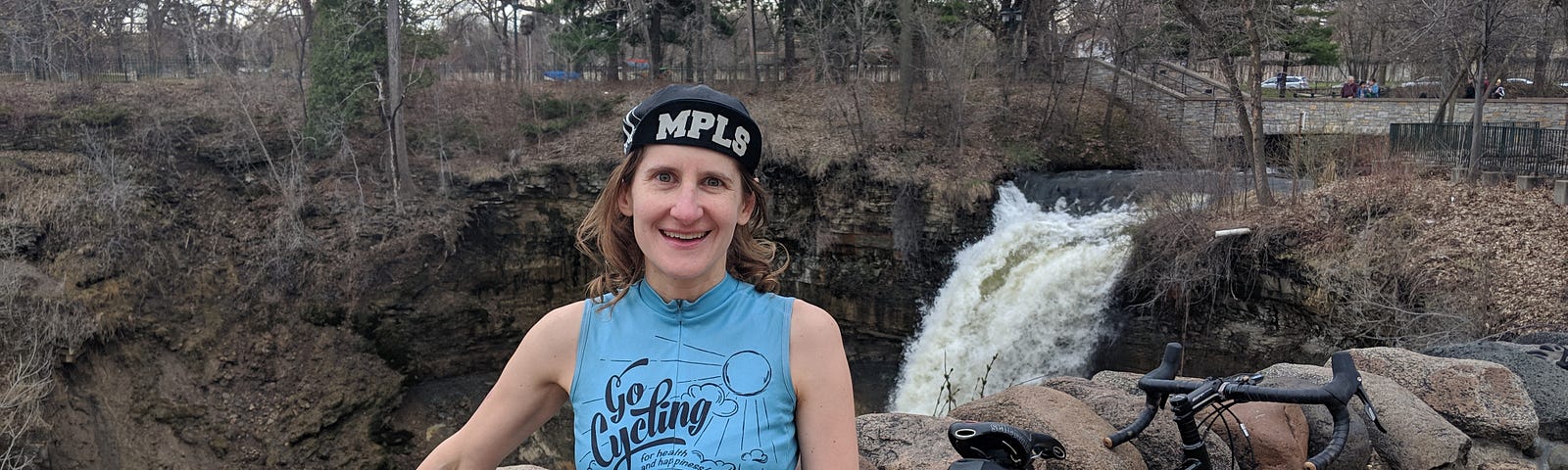 Photo of woman standing with two bicycles in front of a rock retaining wall with a waterfall in the background. Alt text to demonstrate the importance of alt text.