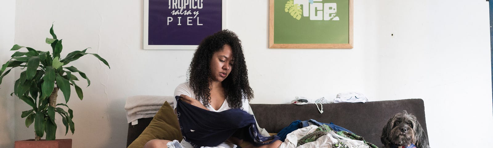 A black woman folds clothes while sitting in her sofa in her living room. There are heaps of clothes.