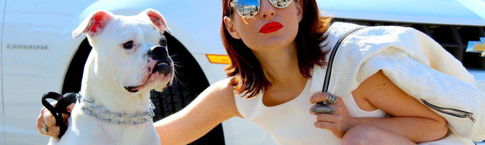 Young brunette in a white t-shirt and shorts and mirrored sunglasses crouched by white bull dog in front of white sports car