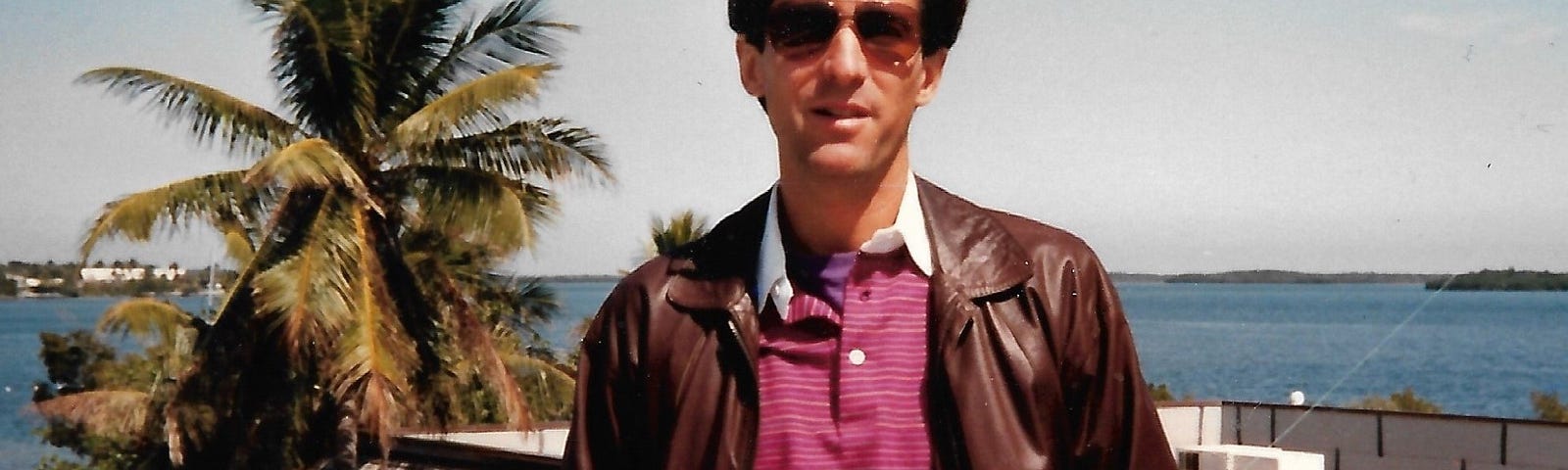 Vintage photo of a light-skinned man with brown hair, wearing aviator shades, a leather jacket, and a polo t-shirt, standing in front of a palm tree and the ocean.