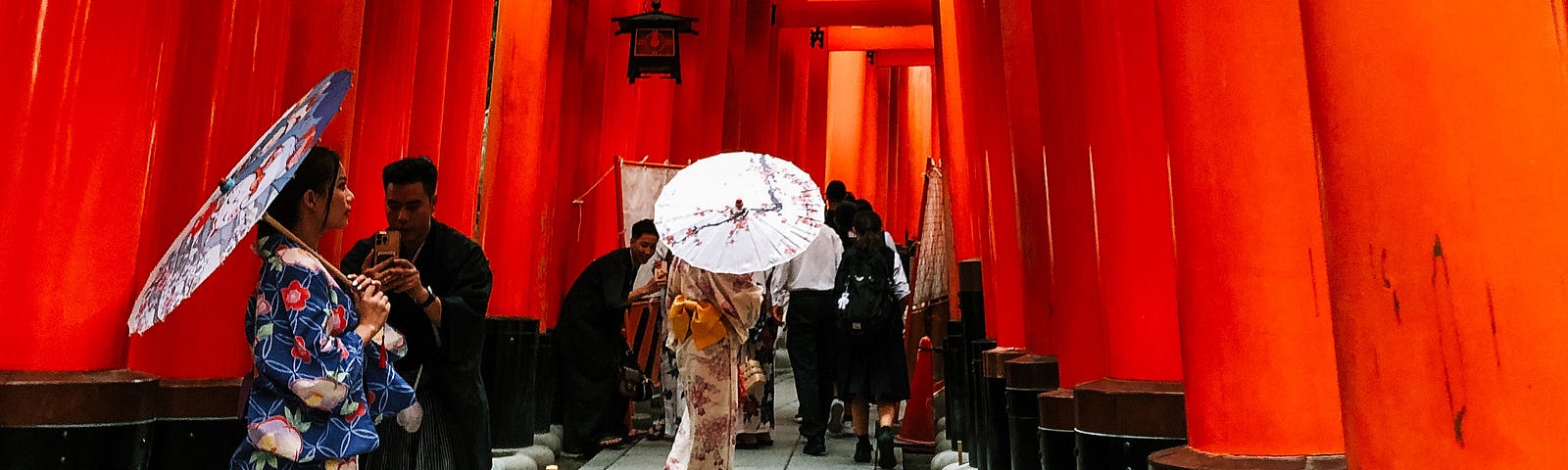 The overwhelmingly red view of Fushimi Inari Shrine in Kyoto, Japan. Two woman are dressed in traditional kimonos with outfit matching umbrellas. A man, also in traditional garb, is taking a photo of one of the women.