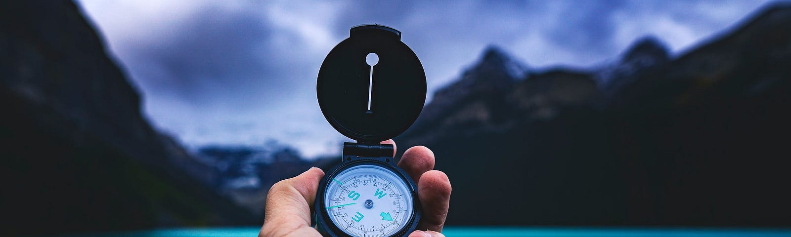 Person Holding A Compass. In the background, it’s clean green colored water and mighty mountains with their shadows in the water. It’s beautiful.