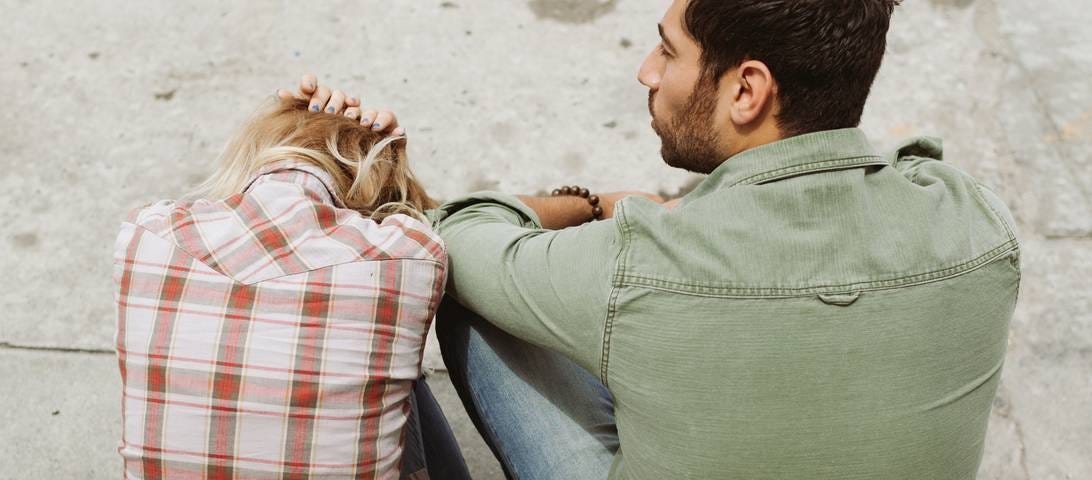 Two people sit on the sidewalk together. One has their head between their knees and their hands on their head. #relationship