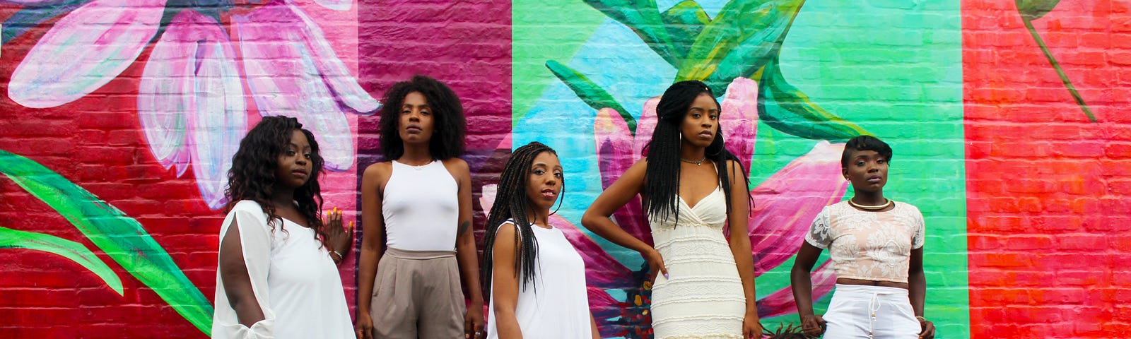A group of African American women wearing various white outfits and posing in front of a painted wall mural.