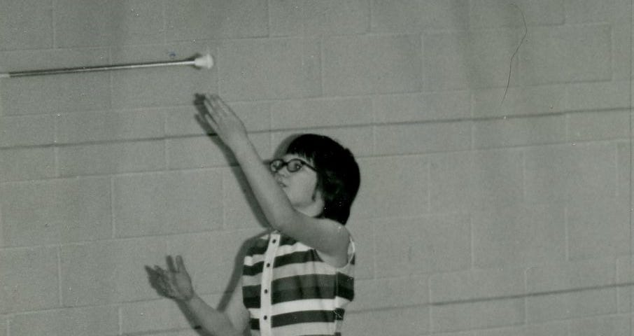 Black and white image from the mid 70’s of a girl in shorts and sleeveless top tossing a baton in the air