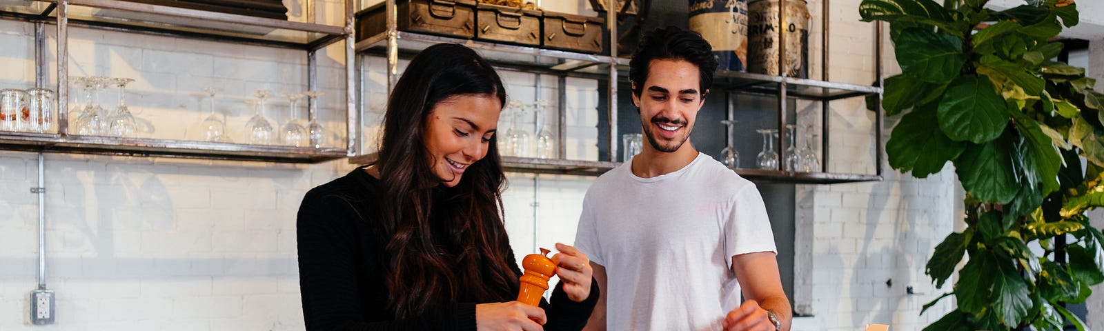 Two smiling young adults in a fancy kitchen. One, probably a woman, is grinding pepper onto a salad.