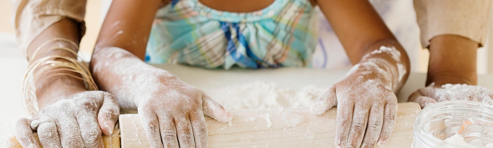 Young Black girl rolling flour with her grandmother, their hands/arms visible on the kitchen top.