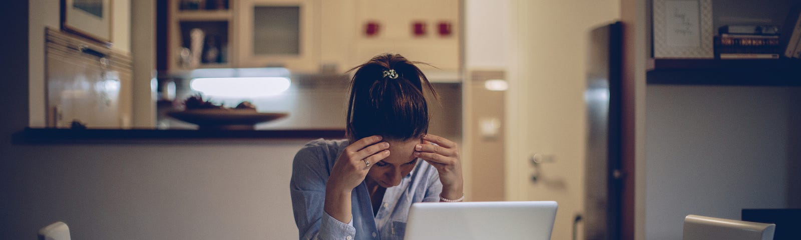 One woman with her head in her hands, sitting alone at home, working on laptop.