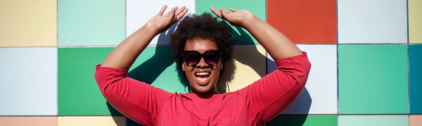 Stylish Black woman in sunglasses and red outfit posing in front of a colorful tiled wall.