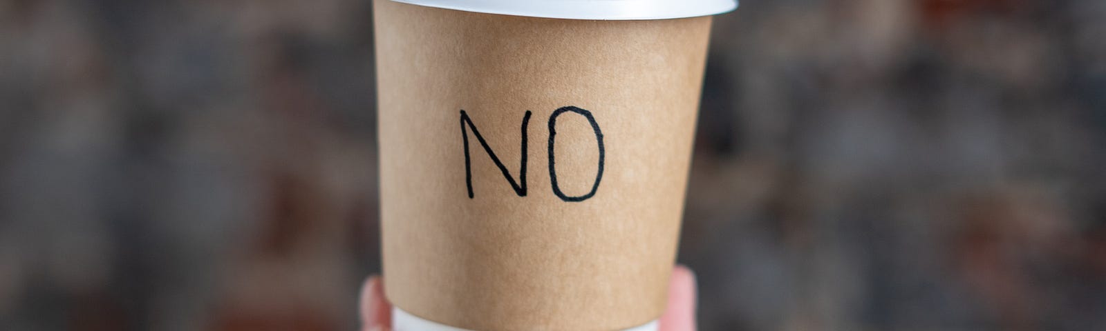 A hand is holding up a disposable coffee cup with the word “NO” written on it.