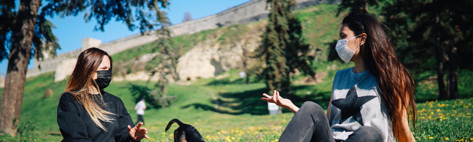 Two young women with protective masks hanging out in the park with a dog during the coronavirus outbreak.