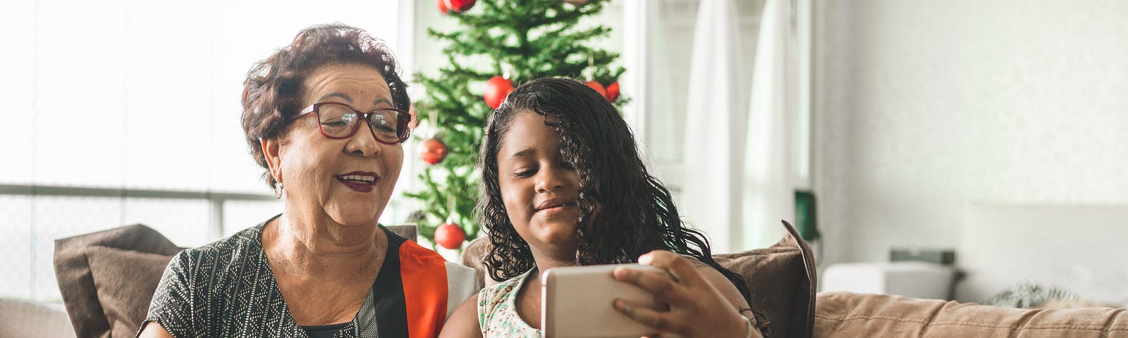 A young girl and her grandmother take a selfie on their couch in front of a Christmas tree.