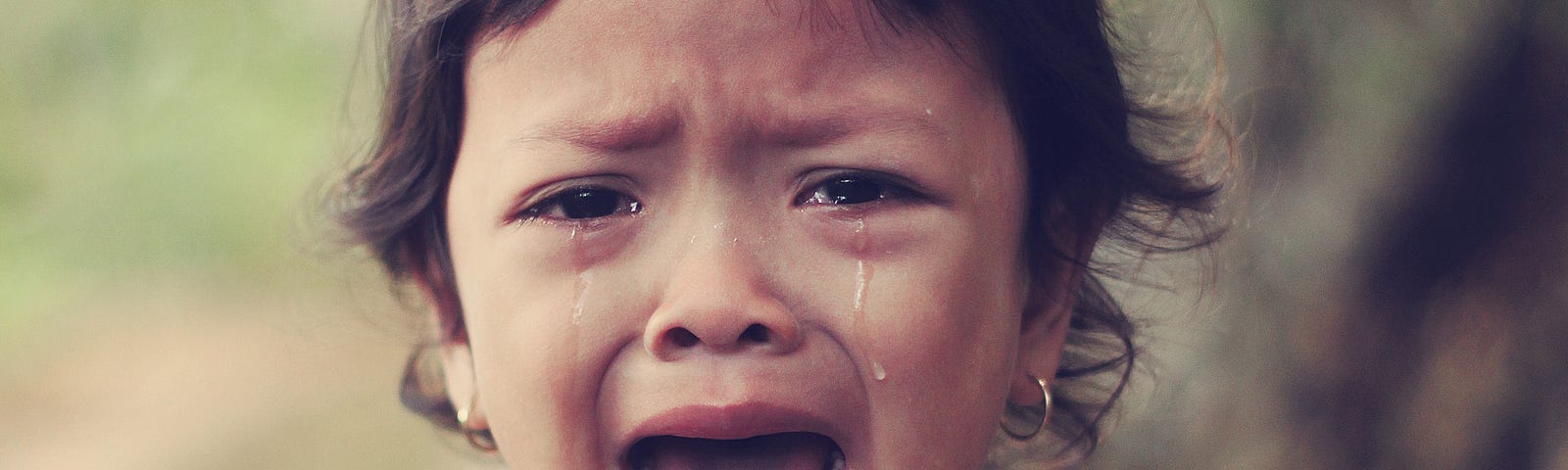 Mouth open and tears running down her cheeks, a toddler in a “happy” tank top cries.