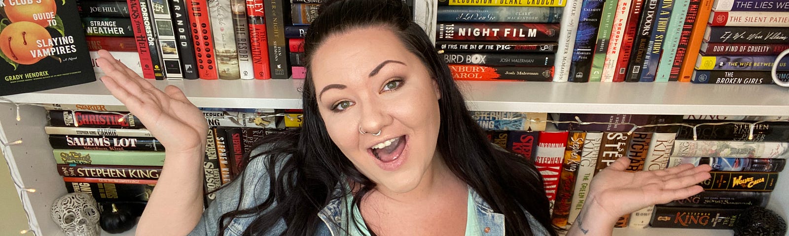 Myself with my hands up, smiling, standing in front of my white bookshelves, full of books.