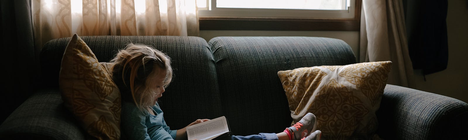 A young child sitting on a couch next to a window, reading a book.