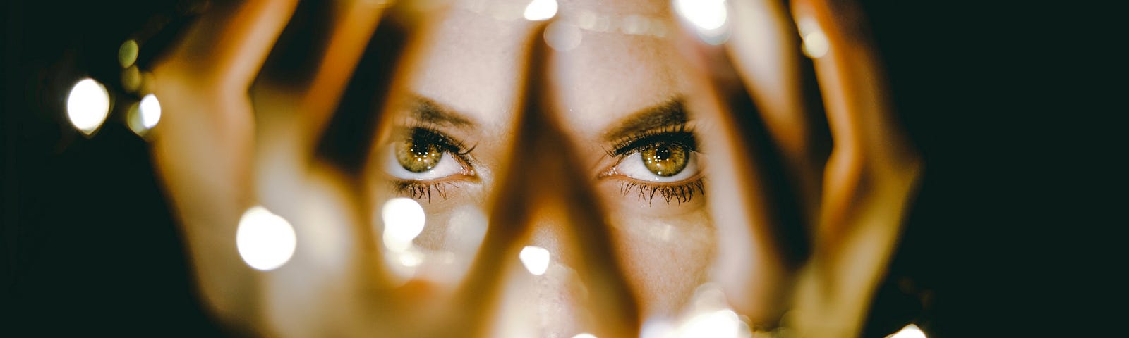 A person’s eyes seen through a gap in the fingers, draped with twinkle lights, they hold in front of their face.