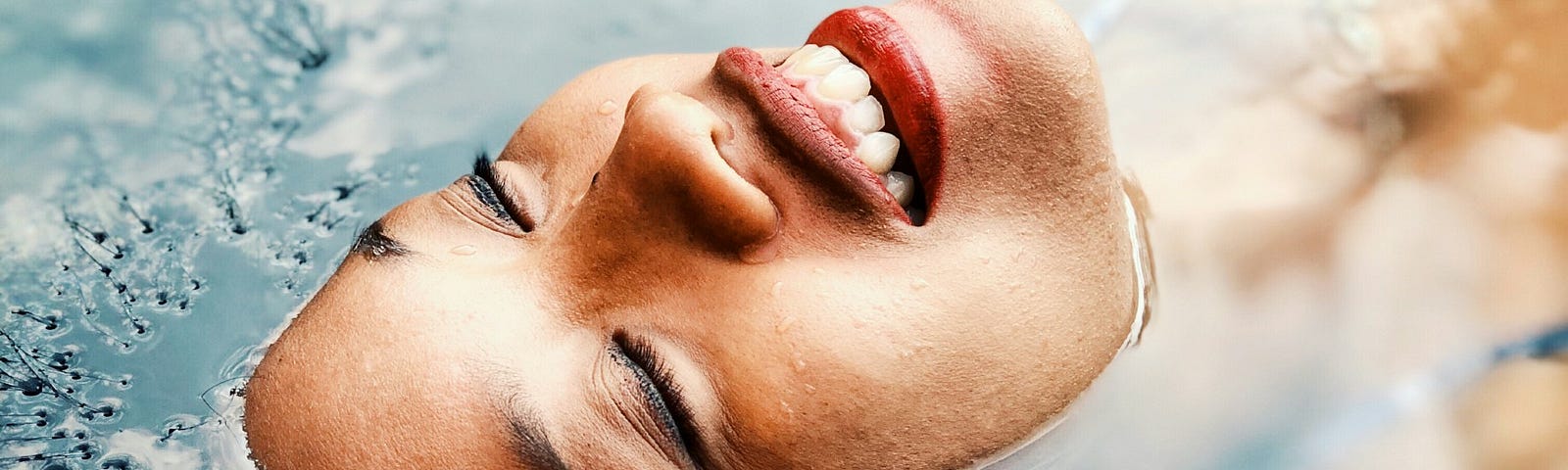 “Embodiment and Reconnecting to Self: Embrace Inner Harmony” This photo shows the face of a woman with carmel-brown skin floating in a pool of blue water. Only her face is above the water and she is smiling.