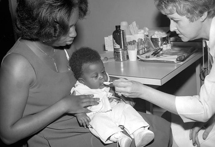 An African American child is receiving an oral vaccine as they are held by their mother.