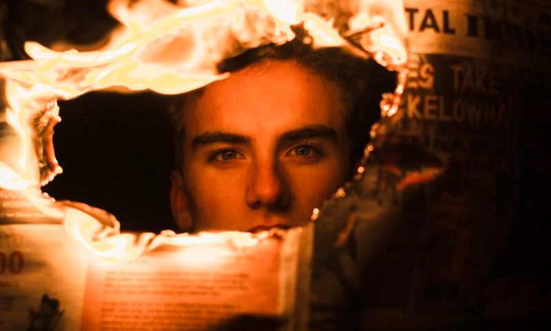 A dark photo of a young man looking through a hole in a burning newspaper.