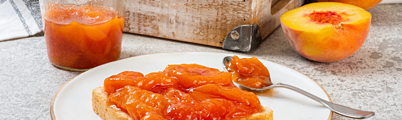 A slice of bread with peach jam on it on a plate in front of a small wooden basket with peaches (whole and halved) in it.