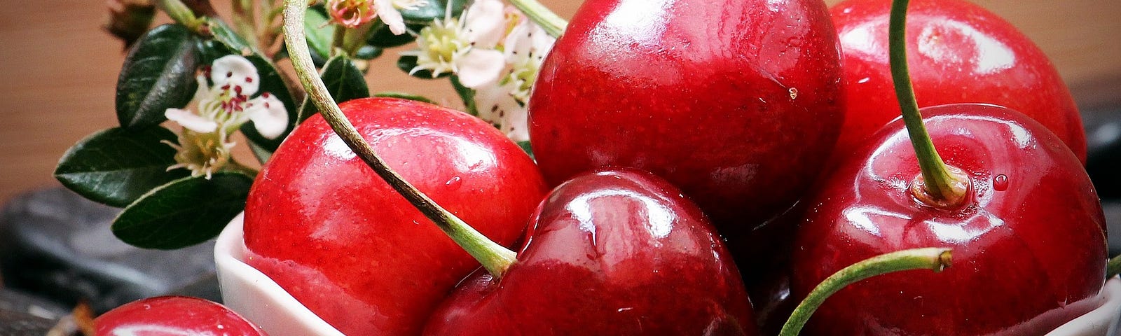 A close-up of a glossy red cherries in a white bowl. A sprig of white blossom and leaves is in the background.