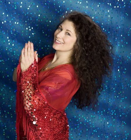 This is a photo of the author standing, smiling, hands together, in red with sparkly blue back drop