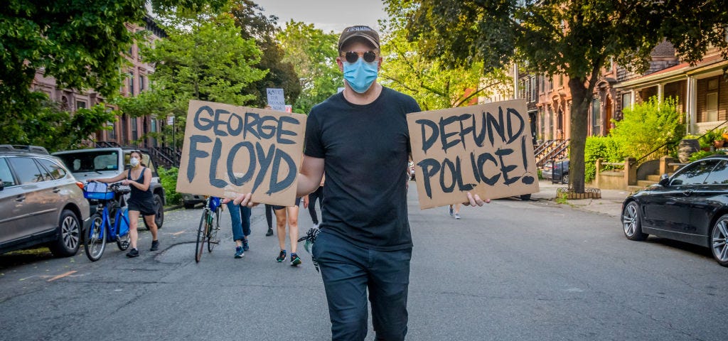 A photo of a protestor holding up two signs that say “GEORGE FLOYD” and “DEFUND POLICE!”