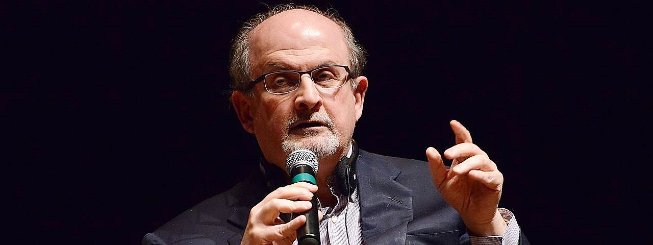 Salman Rushdie making a speech wears a dark suit and raises his finger while holding the microphone against a black background.