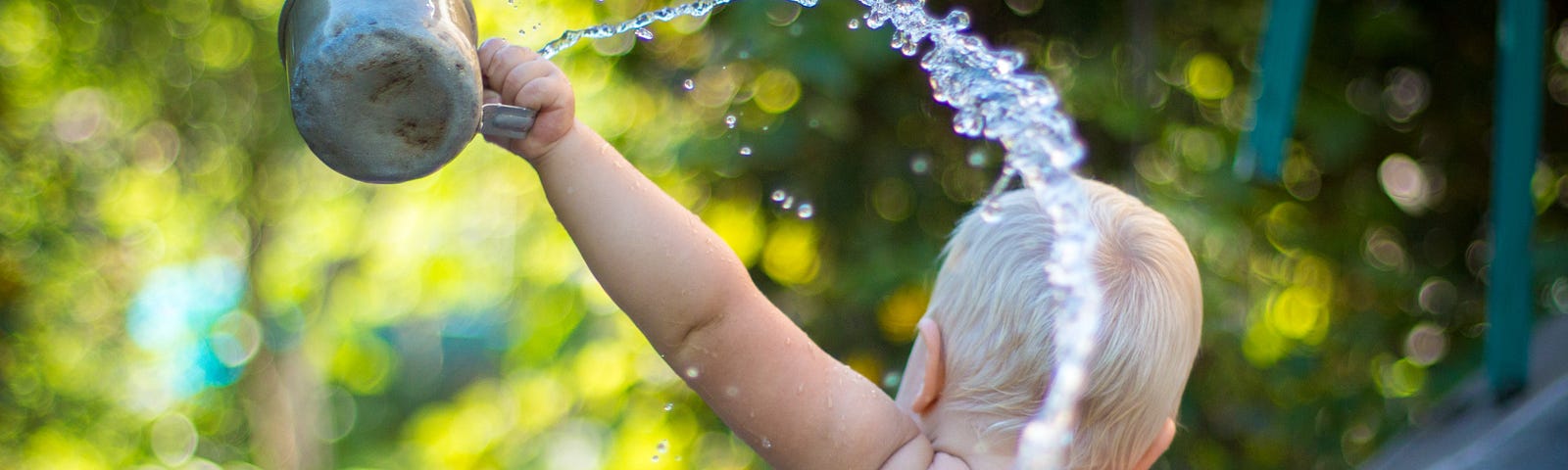 Baby sitting in blue bucket with back to viewer, splashing a big arc of water from an aluminum cup on a soft-focus outdoor watery background.