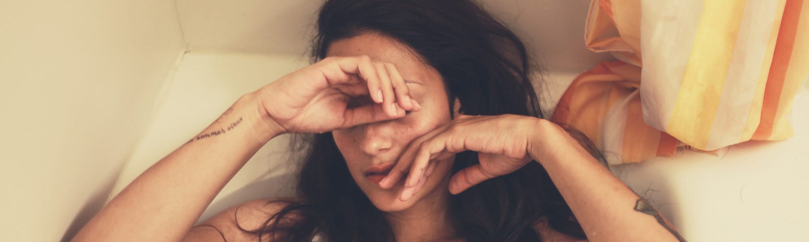 A woman sits in a tub covering her eyes and cheek, seemingly tired.