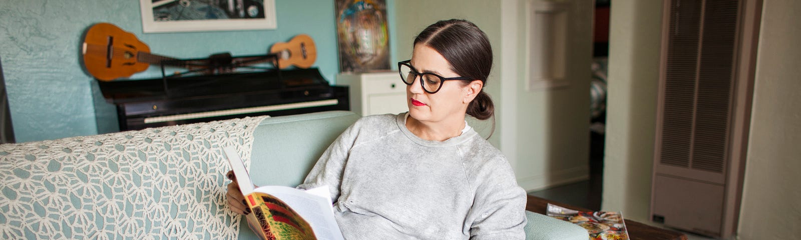 A woman curled up on a blue sofa in her creative home reading a book with her small dog in her lap.