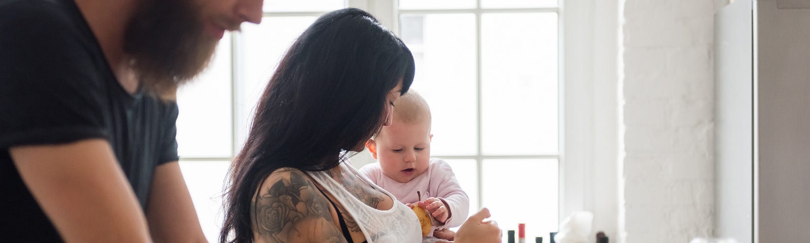 Young mother holding her newborn baby in the kitchen while the father works on his laptop in the foreground.