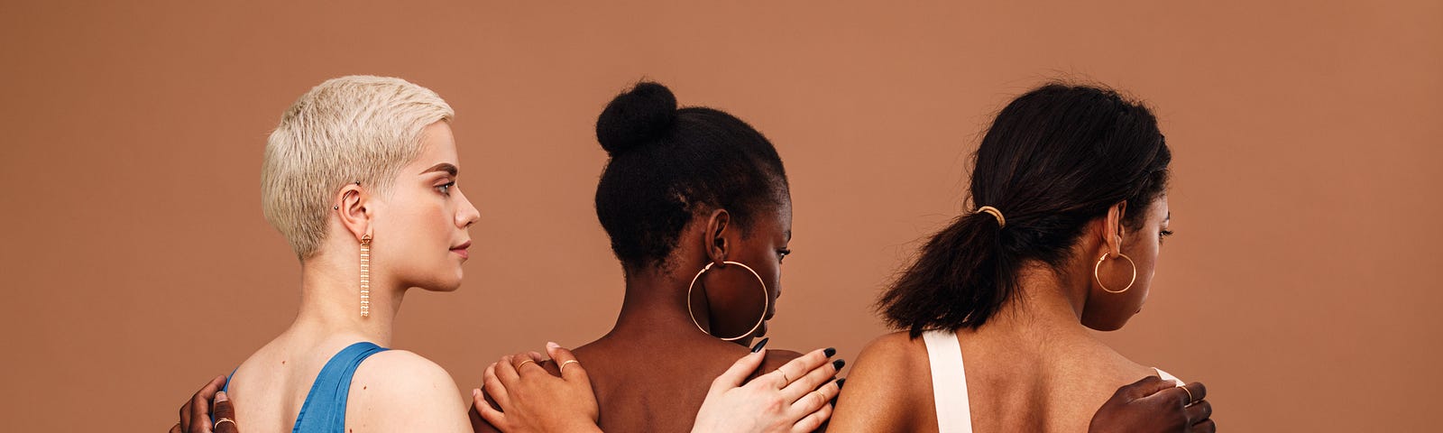 Multi-ethnic women stand with arms on each other’s backs, facing away from the camera, against a light chestnut background.