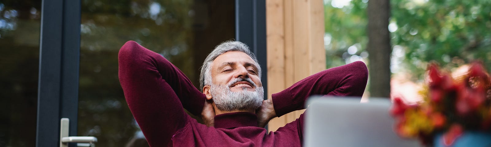 Man with greying beard relaxing outside, hands behind his head, with a relaxed expression.