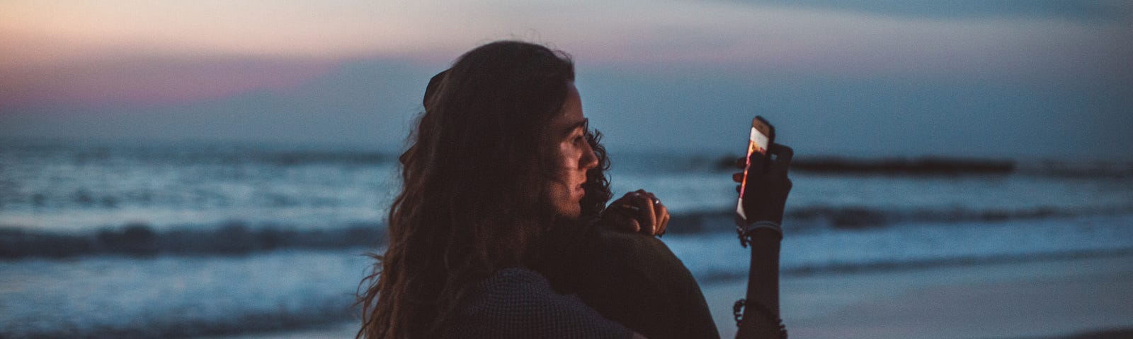 A couple hugs at the beach during sunset while holding their phones out and looking at them.