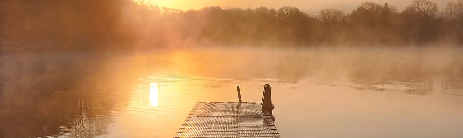 In the middle, a dock extending into the ripples and mists of the water of the lake, the sky is alit by the warm sun on the left rising over the tree line.