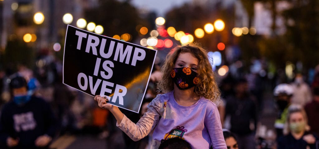 A young girl wearing face mask sits on top of her dad’s shoulders at a protest, holding a sign that says “TRUMP IS OVER.”