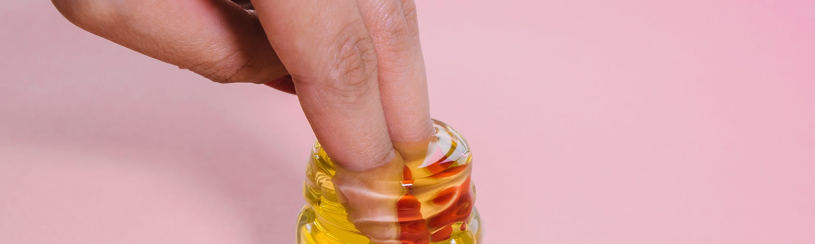 A photo of two fingers dipping in a small jar of honey against a pink background.