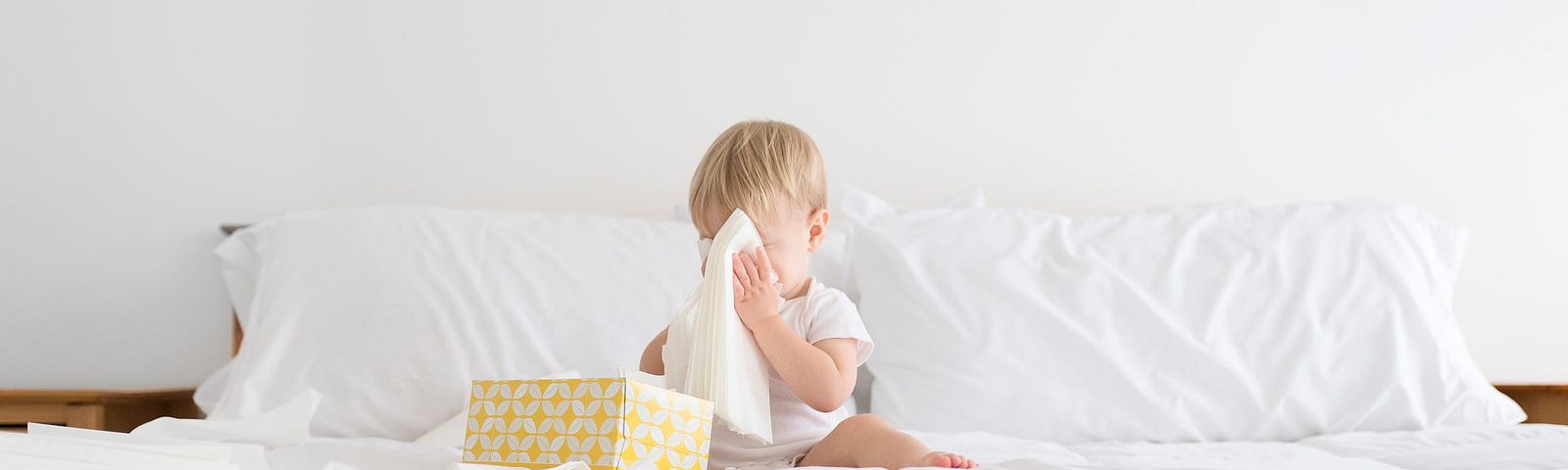 A photo of a baby covering his face with tissues that he’s taken out of a tissue box.